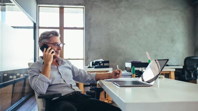Mann sitzt zurückgelehnt am Schreibtisch vor Laptop mit Handy am Ohr Telefonanlage Unternehmen, Telefon