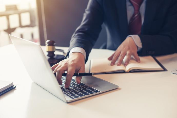 Schreibtisch mit Laptop, Anwalt im Anzug und Stift in der Hand, geöffnetes Buch und Hammer Anwaltskanzlei, IT Anwalt, Kanzleisoftware, Anwalt, Anwalt am Schreibtisch mit Laptop